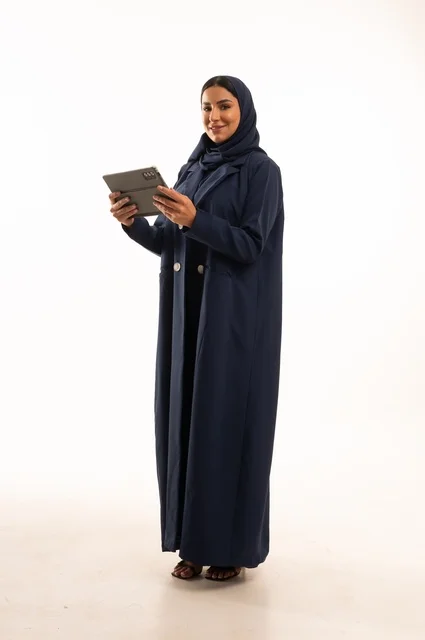 استخدام التقنية الحديثه المتطورة في الحياة العملية عبر الجهاز اللوحي، تعابير وايماءات الاندماج، بورتريه لشابة عربية خليجية سعودية مبتسمة ترتدي الحجاب والعباءة تقوم بالعمل على جهازالتابلت، صورة الجسم بالطول الكامل، خلفية بيضاء
