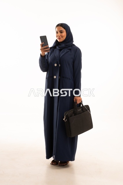 العمل من خلال التقنيات الحديثة متابعة المهام اليومية من خلال الهاتف المحمول, بورتريه لإمرأة عربية خليجية سعودية مبتسمه ترتدي العباءة تحمل حقيبة الأعمال, أناقة النساء الخليجيات, الاهتمام بالمظهر الخارجي, الجسم بالطول الكامل، خلفية بيضاء