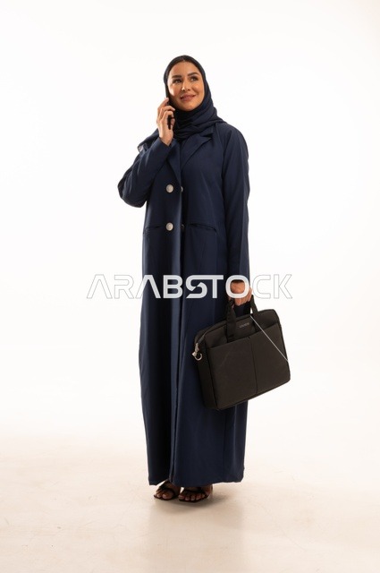 أجراء مكالمة هاتفية العمل من خلال التقنيات الحديثة, بورتريه لإمرأة عربية خليجية سعودية مبتسمه ترتدي العباءة تحمل حقيبة الأعمال, أناقة النساء الخليجيات, الاهتمام بالمظهر الخارجي, الجسم بالطول الكامل، خلفية بيضاء
