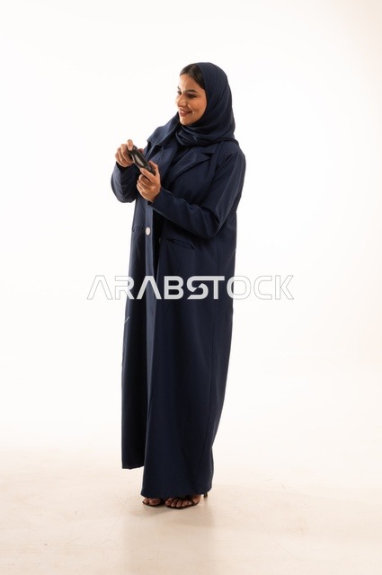 مفهوم الاندماج والتفاعل باللعب ألعاب البلايستيشن، قضاء أوقات ممتعة, بورتريه لشابة عربية خليجية سعودية ترتدي العباءة، سيده سعوديه تمسك بيدها عصا التحكم عن بعد تلعب بألعاب الفيديو، التقنيات الالكترونية الحديثة، الجسم بالطول الكامل، خلفية بيضاء