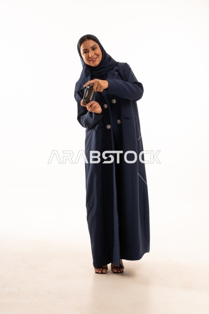 مفهوم الاندماج والتفاعل باللعب ألعاب البلايستيشن، قضاء أوقات ممتعة, بورتريه لشابة عربية خليجية سعودية ترتدي العباءة، سيده سعوديه تمسك بيدها عصا التحكم عن بعد تلعب بألعاب الفيديو، التقنيات الالكترونية الحديثة، الجسم بالطول الكامل، خلفية بيضاء