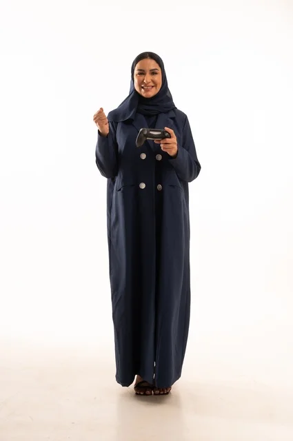 قضاء أوقات ممتعة, بورتريه لشابة عربية خليجية سعودية ترتدي العباءة، سيده سعوديه تمسك بيدها عصا التحكم عن بعد تلعب بألعاب الفيديو بإيماءات وجه ويدين مختلفة تدل على الفوز والإنتصار، الجسم بالطول الكامل، خلفية بيضاء