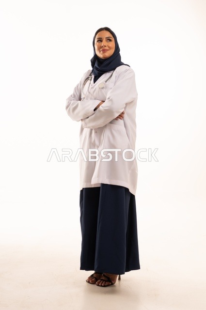 النظر إلى الكاميرا بايماءات الثقة بالنفس تعابير يد متقاطعة، الاهتمام بالمظهر الخارجي، تقديم الفحص الطبي والخدمات العلاجية، بورتريه مقرب لطبيبة عربية خليجية سعودية محجبة مبتسمة ترتدي معطف الطبيب وتضع السماعات الطبية، مفهوم الأنوثة والنعومة، خلفية بيضاء