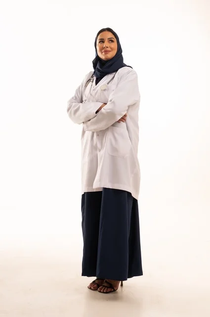 النظر إلى الكاميرا بايماءات الثقة بالنفس تعابير يد متقاطعة، الاهتمام بالمظهر الخارجي، تقديم الفحص الطبي والخدمات العلاجية، بورتريه مقرب لطبيبة عربية خليجية سعودية محجبة مبتسمة ترتدي معطف الطبيب وتضع السماعات الطبية، مفهوم الأنوثة والنعومة، خلفية بيضاء