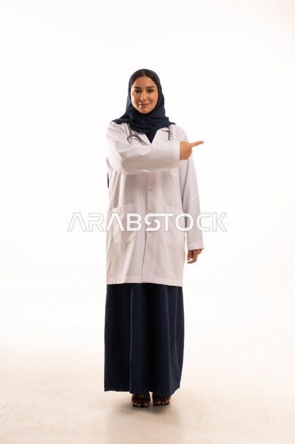 تعابير يد الأتجاه من هذه الجهة ايماءات الثقة بالنفس، الاهتمام بالمظهر الخارجي، تقديم الفحص الطبي والخدمات العلاجية، بورتريه مقرب لطبيبة عربية خليجية سعودية محجبة مبتسمة ترتدي معطف الطبيب وتضع السماعات الطبية، مفهوم الأنوثة والنعومة، خلفية بيضاء