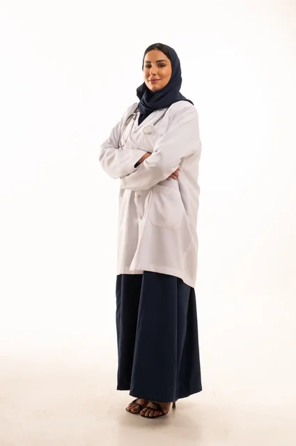 النظر إلى الكاميرا بايماءات الثقة بالنفس تعابير يد متقاطعة، الاهتمام بالمظهر الخارجي، تقديم الفحص الطبي والخدمات العلاجية، بورتريه مقرب لطبيبة عربية خليجية سعودية محجبة مبتسمة ترتدي معطف الطبيب وتضع السماعات الطبية، مفهوم الأنوثة والنعومة، خلفية بيضاء