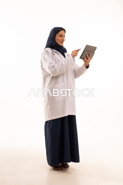استخدام التقنية الحديثه المتطورة في الحياة العملية، متابعة العمل باستخدام جهاز لوحي، بورتريه لإمرأة عربية خليجية سعودية ترتدي الحجاب والعباءة تقوم بالعمل على التابلت، تصفح مواقع التواصل، صورة الجسم بالطول الكامل، خلفية بيضاء