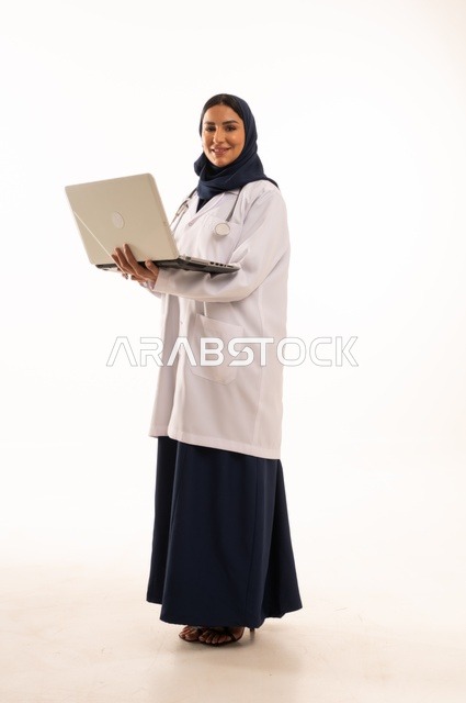 ايماءات وجه السرور، متابعة العمل باستخدام التكنولوجيا ، استخدام التقنية الحديثه المتطورة في الحياة العملية، بورتريه لإمرأة عربية خليجية سعودية ترتدي الحجاب والعباءة تقوم بالعمل على الحاسب المحمول، تصفح مواقع التواصل، صورة الجسم بالطول الكامل، خلفية بيضاء