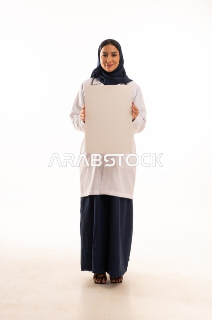 العروض والإعلانات التسويقية, لوحات توضيحية ووسائل تعليمية, بورتريه لطبيبة عربية خليجية سعودية ترتدي العباءة والمئزر الأبيض تمسك بيديها لوحة بيضاء مستطيلة فارغة، شابة سعودية تنظر إلى الكاميرا بتعابير الحزم والجدية، موك اب مستطيل أبيض, خلفية بيضاء