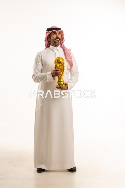 ايماءات السعادة والفرح, مفهوم الربح والفوز، بورتريه لرجل عربي خليجي سعودي يرتدي الثوب التقليدي والشماغ  يحمل بيده كأس العالم الذهبي لرياضة كرة القدم، نشاط بدني للمحافظة على صحة الاجسام و اللياقة، الجسم بالطول الكامل، خلفية بيضاء