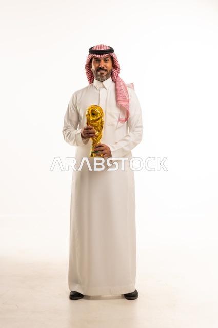 Gestures of happiness and joy, the concept of profit and victory, a portrait of an Arab Gulf Saudi man wearing traditional attire and a ghutrah holding the golden World Cup for football in his hand, physical activity to maintain body health and fitness, full-length body, white background.