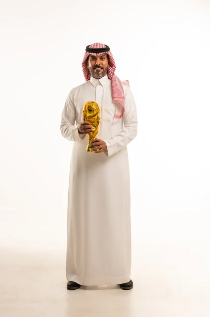 ايماءات السعادة والفرح, مفهوم الربح والفوز، بورتريه لرجل عربي خليجي سعودي يرتدي الثوب التقليدي والشماغ  يحمل بيده كأس العالم الذهبي لرياضة كرة القدم، نشاط بدني للمحافظة على صحة الاجسام و اللياقة، الجسم بالطول الكامل، خلفية بيضاء