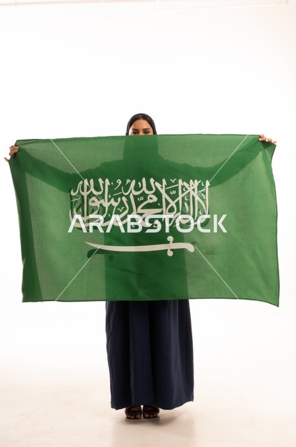 رفع راية التوحيد والإسلام، إحياء ذكرى اليوم الوطني السعودي 23 سبتمبر, النظر بتعابير السرور والفخر, الفخر بالانتماء للوطن, يوم العلم 11 مارس، بورتريه لامرأة عربية خليجية سعودية مبتسمة محجبة ترتدي البالطو تحمل بيدها علم المملكة، الجسم بالطول الكامل, خلفية بيضاء