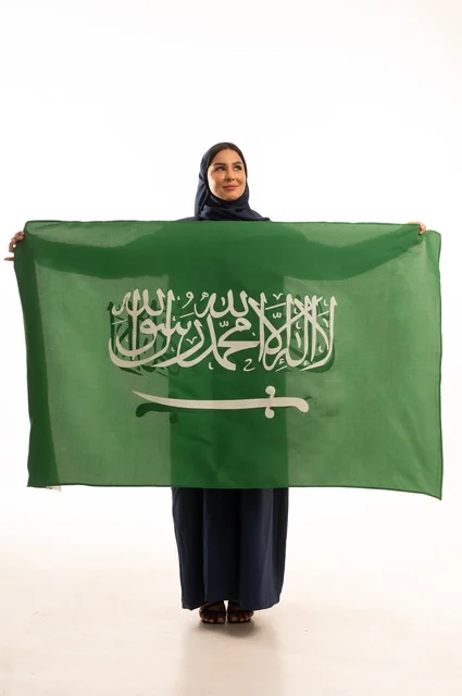 رفع راية التوحيد والإسلام، إحياء ذكرى اليوم الوطني السعودي 23 سبتمبر, النظر بتعابير السرور والفخر, الفخر بالانتماء للوطن, يوم العلم 11 مارس، بورتريه لامرأة عربية خليجية سعودية مبتسمة محجبة ترتدي البالطو تحمل بيدها علم المملكة، الجسم بالطول الكامل, خلفية بيضاء