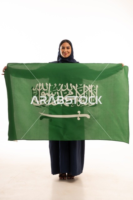 رفع راية التوحيد والإسلام، إحياء ذكرى اليوم الوطني السعودي 23 سبتمبر, النظر بتعابير السرور والفخر, الفخر بالانتماء للوطن, يوم العلم 11 مارس، بورتريه لامرأة عربية خليجية سعودية مبتسمة محجبة ترتدي البالطو تحمل بيدها علم المملكة، الجسم بالطول الكامل, خلفية بيضاء