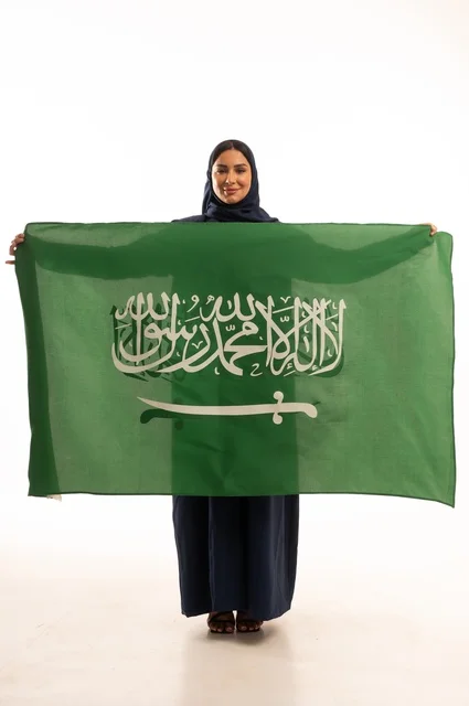 Raising the banner of monotheism and Islam, commemorating the Saudi National Day on September 23, looking with expressions of joy and pride, pride in belonging to the homeland, Flag Day on March 11, a portrait of a smiling Saudi Gulf Arab woman wearing a hijab and an abaya holding the Kingdom's flag in her hand, full-body, white background.