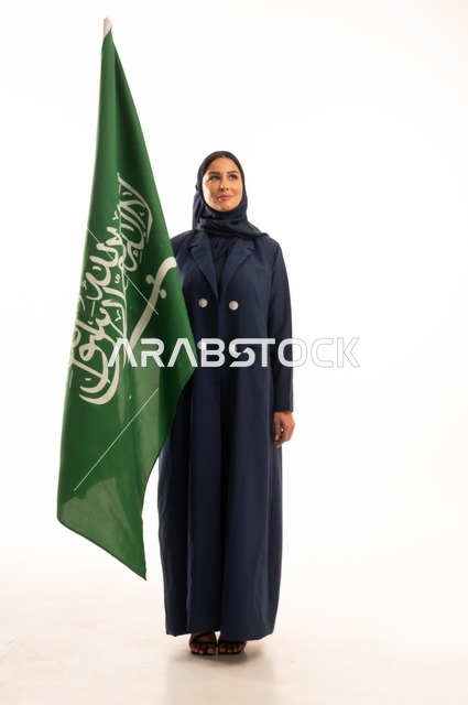 رفع راية التوحيد والإسلام، إحياء ذكرى اليوم الوطني السعودي 23 سبتمبر, النظر بتعابير السرور والفخر, الفخر بالانتماء للوطن, يوم العلم 11 مارس، بورتريه لامرأة عربية خليجية سعودية مبتسمة محجبة ترتدي البالطو تحمل بيدها علم المملكة، الجسم بالطول الكامل, خلفية بيضاء