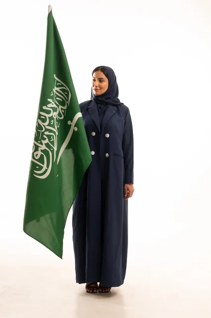 رفع راية التوحيد والإسلام، إحياء ذكرى اليوم الوطني السعودي 23 سبتمبر, النظر بتعابير السرور والفخر, الفخر بالانتماء للوطن, يوم العلم 11 مارس، بورتريه لامرأة عربية خليجية سعودية مبتسمة محجبة ترتدي البالطو تحمل بيدها علم المملكة، الجسم بالطول الكامل, خلفية بيضاء