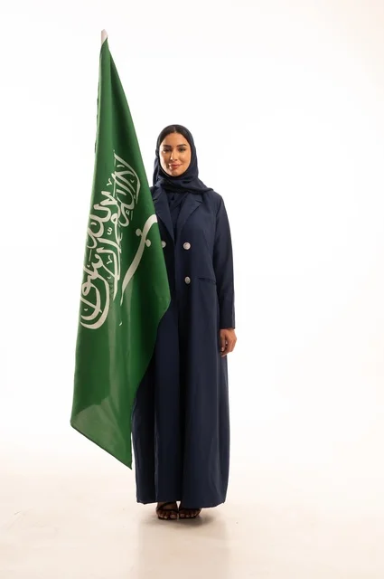 رفع راية التوحيد والإسلام، إحياء ذكرى اليوم الوطني السعودي 23 سبتمبر, النظر بتعابير السرور والفخر, الفخر بالانتماء للوطن, يوم العلم 11 مارس، بورتريه لامرأة عربية خليجية سعودية مبتسمة محجبة ترتدي البالطو تحمل بيدها علم المملكة، الجسم بالطول الكامل, خلفية بيضاء