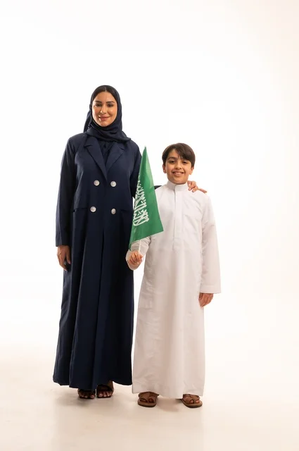 Raising children to be proud of their belonging to the homeland, raising the banner of monotheism and Islam, commemorating the Saudi National Day on September 23, the Flag Day on March 11, a portrait of a smiling Saudi Gulf Arab woman wearing a hijab and a coat standing with her son holding the Kingdom's flag in her hand, full-body, white background.