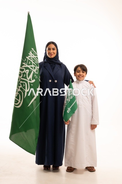 تربية الابناء على الفخر بالانتماء للوطن, رفع راية التوحيد والإسلام، إحياء ذكرى اليوم الوطني السعودي 23 سبتمبر,  يوم العلم 11 مارس، بورتريه لامرأة عربية خليجية سعودية مبتسمة محجبة ترتدي البالطو تقف مع ابنها تحمل بيدها علم المملكة، الجسم بالطول الكامل، خلفية بيضاء