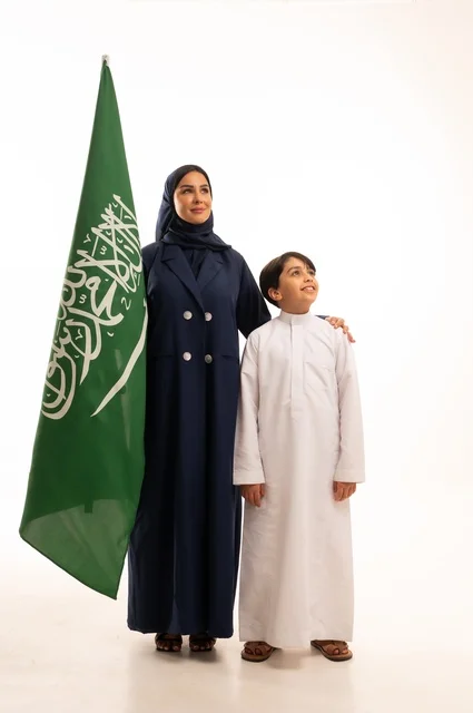 تربية الابناء على الفخر بالانتماء للوطن, رفع راية التوحيد والإسلام، إحياء ذكرى اليوم الوطني السعودي 23 سبتمبر,  يوم العلم 11 مارس، بورتريه لامرأة عربية خليجية سعودية مبتسمة محجبة ترتدي البالطو تقف مع ابنها تحمل بيدها علم المملكة، الجسم بالطول الكامل، خلفية بيضاء