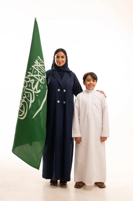 تربية الابناء على الفخر بالانتماء للوطن, رفع راية التوحيد والإسلام، إحياء ذكرى اليوم الوطني السعودي 23 سبتمبر,  يوم العلم 11 مارس، بورتريه لامرأة عربية خليجية سعودية مبتسمة محجبة ترتدي البالطو تقف مع ابنها تحمل بيدها علم المملكة، الجسم بالطول الكامل، خلفية بيضاء
