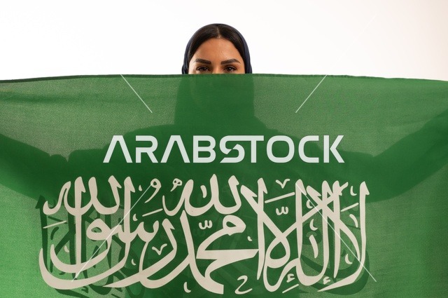 Pride in belonging to the homeland, raising the banner of monotheism and Islam, commemorating the Saudi National Day on September 23, looking at the camera with expressions of joy, Flag Day on March 11, a close-up portrait of a smiling Saudi Gulf Arab woman wearing a hijab and an abaya holding the Kingdom's flag in her hand, with a white background.