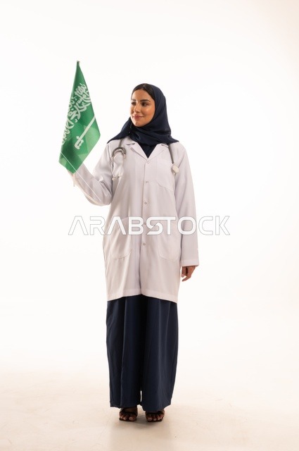 تقديم الخدمات الصحية للمرضى، إحياء ذكرى اليوم الوطني السعودي 23 سبتمبر، مفهوم الطب والرعاية الصحية، بورتريه لطبيبة عربية خليجية سعودية محجبة ترتدي المعطف والسماعة الطبية تحمل بيدها علم المملكة، العمل في القطاع الصحي في المملكة، الجسم بالطول الكامل، خلفيه بيضاء