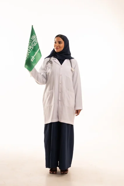 Providing health services to patients, commemorating Saudi National Day on September 23, the concept of medicine and healthcare, a portrait of a Saudi Gulf Arab female doctor wearing a hijab, a coat, and a stethoscope holding the Kingdom's flag in her hand, working in the health sector in the Kingdom, full-body, white background.