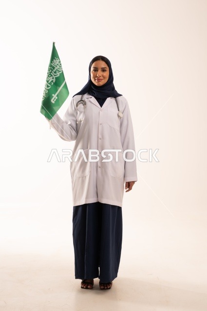 تقديم الخدمات الصحية للمرضى، إحياء ذكرى اليوم الوطني السعودي 23 سبتمبر، مفهوم الطب والرعاية الصحية، بورتريه لطبيبة عربية خليجية سعودية محجبة ترتدي المعطف والسماعة الطبية تحمل بيدها علم المملكة، العمل في القطاع الصحي في المملكة، الجسم بالطول الكامل، خلفيه بيضاء