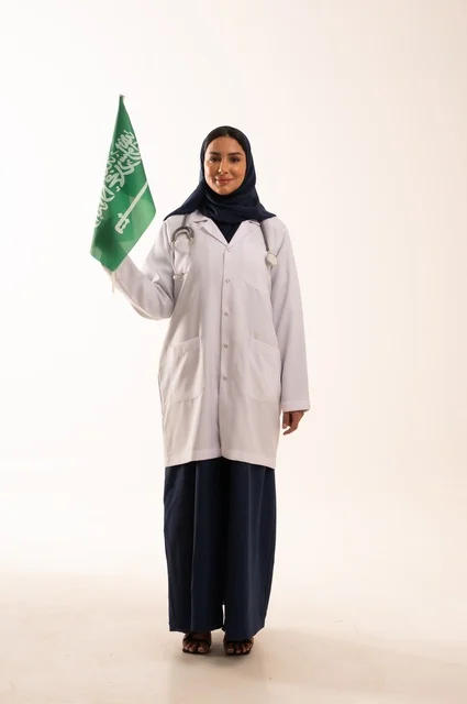 تقديم الخدمات الصحية للمرضى، إحياء ذكرى اليوم الوطني السعودي 23 سبتمبر، مفهوم الطب والرعاية الصحية، بورتريه لطبيبة عربية خليجية سعودية محجبة ترتدي المعطف والسماعة الطبية تحمل بيدها علم المملكة، العمل في القطاع الصحي في المملكة، الجسم بالطول الكامل، خلفيه بيضاء