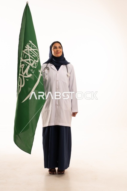 Providing health services to patients, commemorating Saudi National Day on September 23, the concept of medicine and healthcare, a portrait of a Saudi Gulf Arab female doctor wearing a hijab, a coat, and a stethoscope holding the Kingdom's flag in her hand, working in the health sector in the Kingdom, full-body, white background.