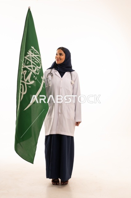 تقديم الخدمات الصحية للمرضى، إحياء ذكرى اليوم الوطني السعودي 23 سبتمبر، مفهوم الطب والرعاية الصحية، بورتريه لطبيبة عربية خليجية سعودية محجبة ترتدي المعطف والسماعة الطبية تحمل بيدها علم المملكة، العمل في القطاع الصحي في المملكة، الجسم بالطول الكامل، خلفيه بيضاء