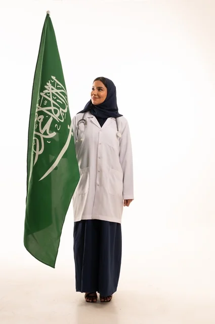 Providing health services to patients, commemorating Saudi National Day on September 23, the concept of medicine and healthcare, a portrait of a Saudi Gulf Arab female doctor wearing a hijab, a coat, and a stethoscope holding the Kingdom's flag in her hand, working in the health sector in the Kingdom, full-body, white background.