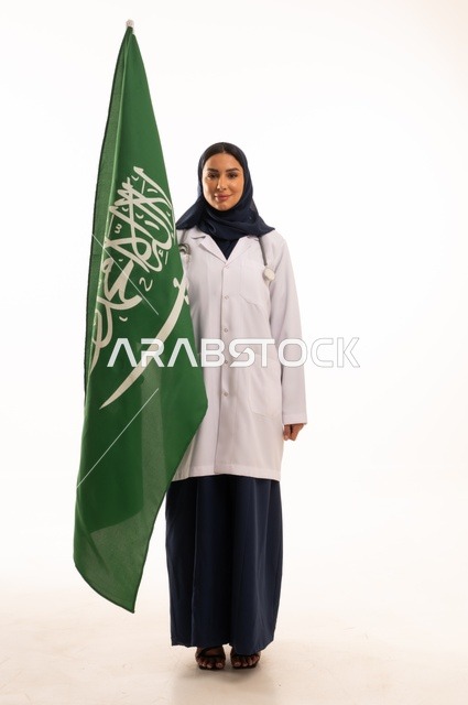 تقديم الخدمات الصحية للمرضى، إحياء ذكرى اليوم الوطني السعودي 23 سبتمبر، مفهوم الطب والرعاية الصحية، بورتريه لطبيبة عربية خليجية سعودية محجبة ترتدي المعطف والسماعة الطبية تحمل بيدها علم المملكة، العمل في القطاع الصحي في المملكة، الجسم بالطول الكامل، خلفيه بيضاء
