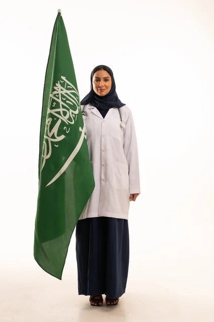 تقديم الخدمات الصحية للمرضى، إحياء ذكرى اليوم الوطني السعودي 23 سبتمبر، مفهوم الطب والرعاية الصحية، بورتريه لطبيبة عربية خليجية سعودية محجبة ترتدي المعطف والسماعة الطبية تحمل بيدها علم المملكة، العمل في القطاع الصحي في المملكة، الجسم بالطول الكامل، خلفيه بيضاء