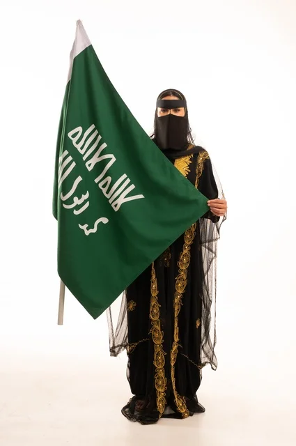 The establishment of the first Saudi state on February 22, the anniversary of our beginning in 1727 AD, wearing the traditional attire of the central region during popular and national occasions, a portrait of an Arab Gulf Saudi woman wearing the burqa and shayla holding the flag and emblem of the Foundation Day with expressions of pride and honor, full-body, white background.