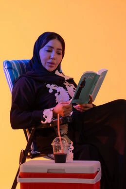 بورتريه لامراه عربيه خليجيه اماراتيه ترتدي العباءه المطرزة والحجاب تجلس على كرسي , قضاء وقت ممتع في فصل الصيف, مفهوم القراءه والثقافة ، شرب المشروبات الصيفية المنعشة ، سيدة سعودية تمسك بيدها كتاب ،  خلفية صفراء
