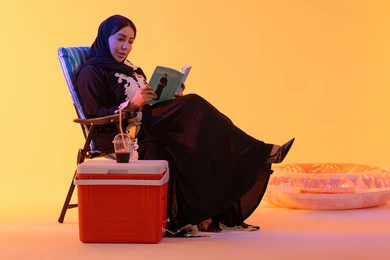 Drinking refreshing summer drinks, having a good time in the summer, the concept of reading and culture, a portrait of an Emirati Gulf Arab woman wearing an embroidered abaya and hijab sitting on a chair, a Saudi woman holding a book in her hand, yellow background.
