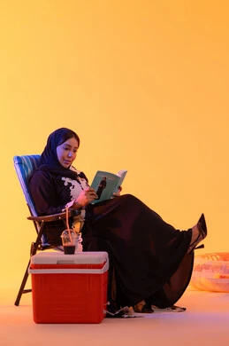 The concept of reading and culture, spending enjoyable time in the summer, drinking refreshing summer beverages, a portrait of an Arab Gulf Emirati woman wearing an embroidered abaya and hijab sitting on a chair, a Saudi woman holding a book in her hand, yellow background.