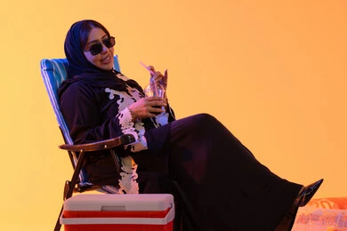 سيدة سعودية تمسك بيدها حافظة البرودة، قضاء وقت ممتع في فصل الصيف, شرب المشروبات الصيفية المنعشة ، بورتريه لامراه عربيه خليجيه اماراتيه ترتدي العباءه المطرزة والحجاب تجلس على كرسي ,  خلفية صفراء