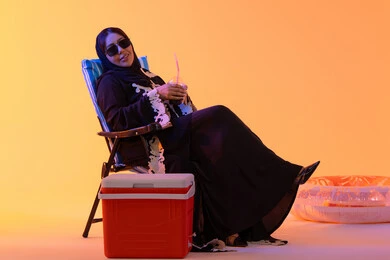 شرب المشروبات الصيفية المنعشة ،قضاء وقت ممتع في فصل الصيف,  بورتريه لامراه عربيه خليجيه اماراتيه ترتدي العباءه المطرزة والحجاب تجلس على كرسي , سيدة سعودية تمسك بيدها حافظة البرودة،  خلفية صفراء