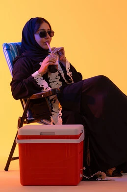 قضاء وقت ممتع في فصل الصيف, شرب المشروبات الصيفية المنعشة ، بورتريه لامراه عربيه خليجيه اماراتيه ترتدي العباءه المطرزة والحجاب تجلس على كرسي , سيدة سعودية تمسك بيدها حافظة البرودة،  خلفية صفراء