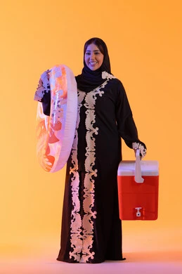A portrait of an Emirati Gulf Arab woman wearing an embroidered abaya and hijab, holding a float in preparation for swimming at the beach. A Saudi lady is holding a cooler in her hand, enjoying a good time in the summer, with a yellow background.