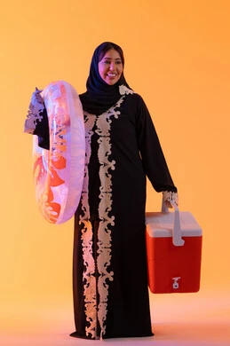 Spending a good time in the summer, a portrait of an Emirati Gulf Arab woman wearing an embroidered abaya and hijab, and a float ready for swimming at the beach, a Saudi woman holding a cooler in her hand, yellow background.