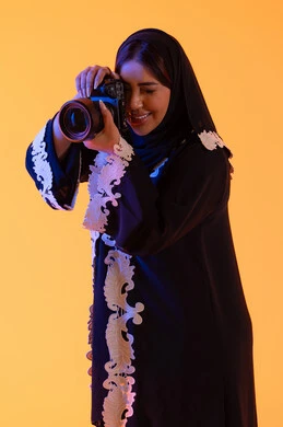 High-definition camera, the concept of professional photography, a Saudi woman practicing her favorite hobbies, capturing memorable photos, a portrait of an Arab Gulf Emirati woman holding a high-definition camera in her hand, an Emirati photographer, yellow background.