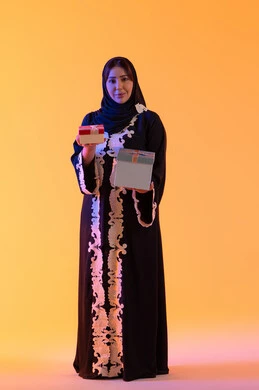The concept of femininity and softness, holidays and happy occasions, celebration and gift-giving, a portrait of an Emirati Gulf Arab woman wearing a hijab and abaya holding a wrapped gift box in her hand, a Saudi woman standing with gestures of happiness and joy, attention to beauty, elegance, and appearance, yellow background.