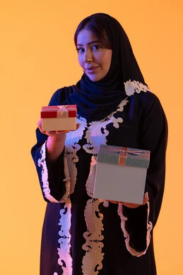 بورتريه لامرأة عربية خليجية اماراتيه ترتدي الحجاب والعباءة تحمل بيدها علبة هدية مغلفة ، سيده سعوديه تقف بايماءات السعادة والسرور، مفهوم الأنوثة والنعومة، الاهتمام بالجمال والأناقة والمظهر الخارجي، الأعياد والمناسبات السعيدة، الاحتفال وتقديم الهدايا،خلفية صفراء