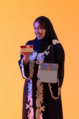 Holidays and happy occasions, celebration and gift-giving, a portrait of an Emirati Gulf Arab woman wearing a hijab and abaya holding a wrapped gift box in her hand, a Saudi woman standing with gestures of happiness and joy, the concept of femininity and softness, attention to beauty, elegance, and appearance, yellow background.